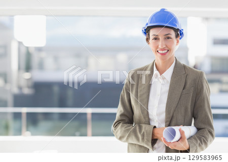 Woman holding plans wearing blazer, blue safety helmet on balcony with glass railing, copy space 129583965