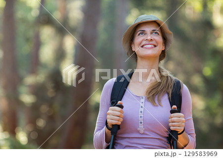 Female hiker standing on forest trail holding poles wearing lavender top bucket hat and backpack 129583969
