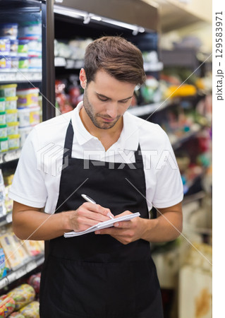 Man in black apron writing in spiral notepad with pen by refrigerated shelves in supermarket aisle Man in black apron writing in spiral notepad with pen by refrigerated shelves in supermarket aisle 129583971