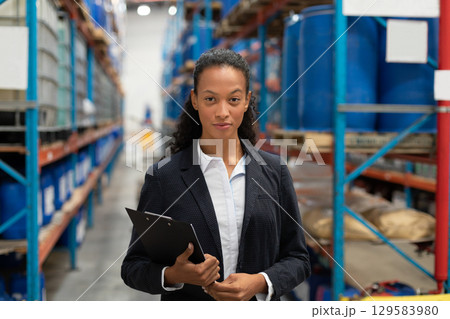 African American businesswoman standing in warehouse aisle reviewing clipboard near metal racks 129583980
