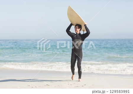 Male surfer wearing wetsuit lifting surfboard overhead on sandy shore under clear sky, copy space 129583991
