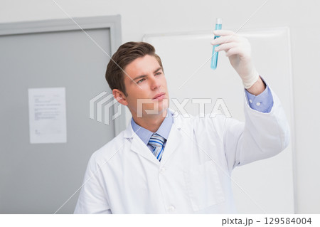 Male laboratory scientist wearing lab coat and latex gloves inspecting blue liquid test tube in lab 129584004