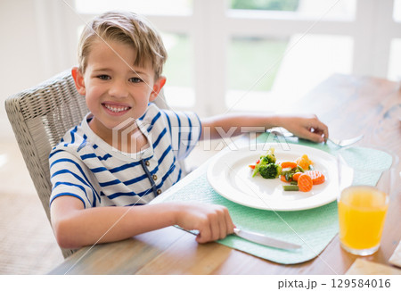 Child eating steamed vegetables with fork and knife and drinking orange juice at home dining table 129584016