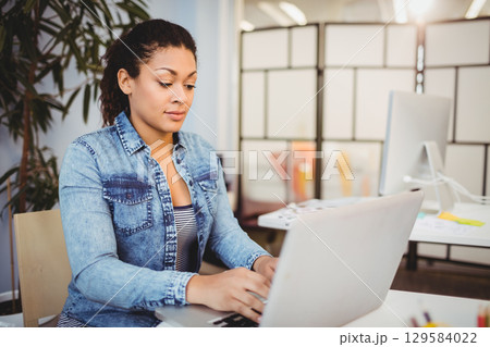 African American woman typing on laptop at office desk with monitor, partition screen, potted plant 129584022