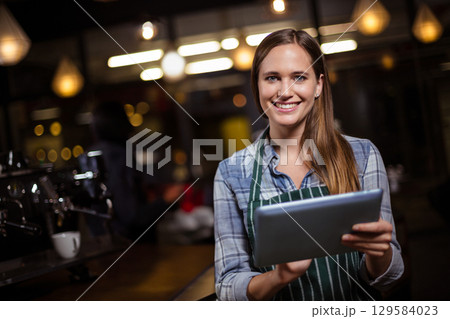 Female barista standing behind wooden counter in cafe using tablet by espresso machine, copy space 129584023