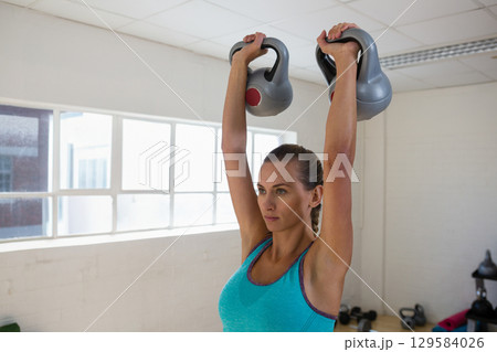 Fit woman lifting two gray kettlebells overhead in fitness room by window wearing teal tank top Fit woman lifting two gray kettlebells overhead in fitness room by window wearing teal tank top 129584026