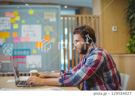 Man wearing plaid shirt and headphones working on laptop at office near sticky notes, copy space Man wearing plaid shirt and headphones working on laptop at office near sticky notes, copy space 129584027