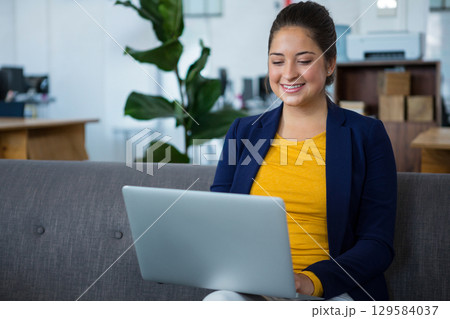 Woman working on laptop while sitting on gray sofa in modern office lounge wearing business attire Woman working on laptop while sitting on gray sofa in modern office lounge wearing business attire 129584037