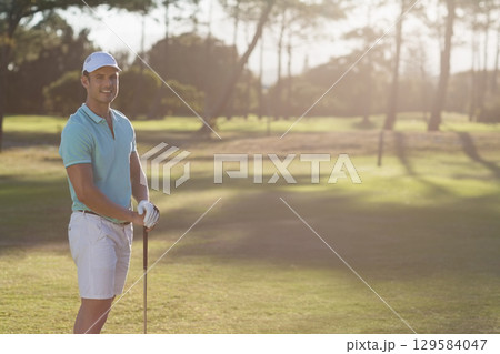 Smiling man wearing glove holding golf club on golf course fairway in warm sunlight, copy space 129584047