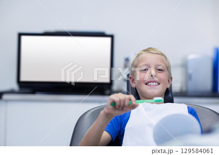Smiling boy reclining in dental chair holding toothbrush near dental mirror at clinic, copy space Smiling boy reclining in dental chair holding toothbrush near dental mirror at clinic, copy space 129584061