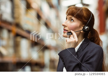Businesswoman in blazer checking stock with headset in warehouse near racks of boxes, copy space 129584065