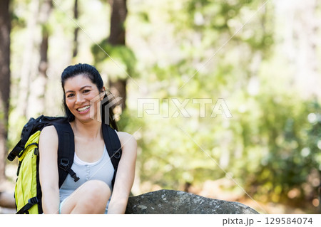 Mature Asian woman sitting on rock in forest wearing yellow and black hiking backpack, smiling Mature Asian woman sitting on rock in forest wearing yellow and black hiking backpack, smiling 129584074