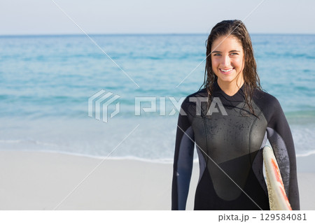 Female surfer standing at water's edge wearing wetsuit and holding surfboard at beach, copy space 129584081