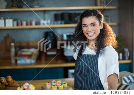 Smiling African American woman serving cupcakes behind counter in café wearing apron, copy space 129584082
