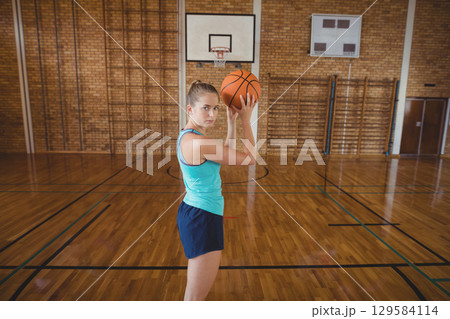 Female athlete in sportswear holding basketball and practicing jump shot on court under backboard 129584114
