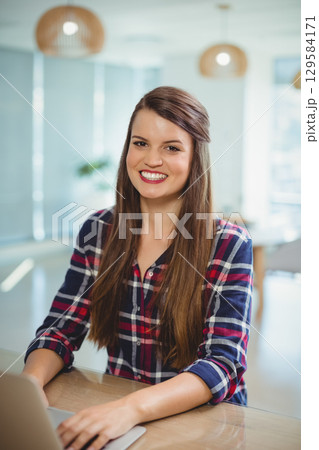 Female office worker smiling while typing on laptop at wood desk in office with wooden lamps 129584171