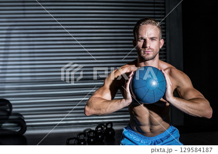 Shirtless man squatting in gym gripping blue medicine ball above black kettlebells, copy space Shirtless man squatting in gym gripping blue medicine ball above black kettlebells, copy space 129584187