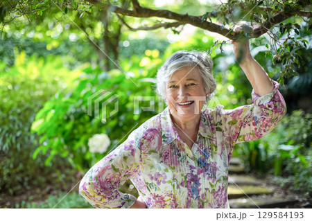 Senior woman standing in garden holding tree branch and wearing floral-print blouse on stone path Senior woman standing in garden holding tree branch and wearing floral-print blouse on stone path 129584193