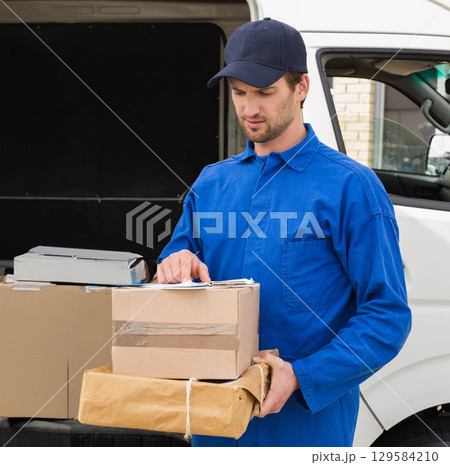 Male courier in uniform reviewing clipboard and holding parcels beside white van on paved driveway 129584210