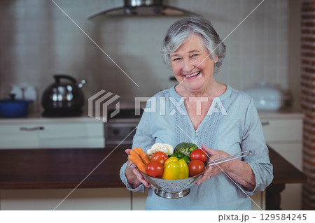Senior woman smiling while holding metal colander of fresh vegetables in home kitchen Senior woman smiling while holding metal colander of fresh vegetables in home kitchen 129584245