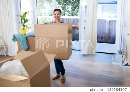 Mid adult Asian woman carrying cardboard box inside bright living room with bubble wrap Mid adult Asian woman carrying cardboard box inside bright living room with bubble wrap 129584507