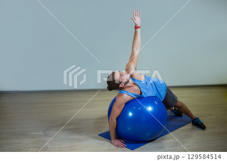 Man wearing blue top and shorts stretching sideways over blue stability ball on mat in gym Man wearing blue top and shorts stretching sideways over blue stability ball on mat in gym 129584514