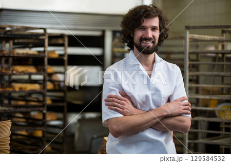 Mid adult man baker crossing arms and smiling in bakery area with metal racks of bread 129584532