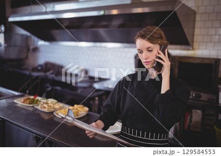 Female chef in chef coat holding smartphone reviewing order sheet at counter in kitchen, copy space 129584553