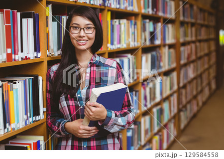 Asian woman in plaid shirt leaning against bookshelf holding blue book in library aisle, copy space 129584558