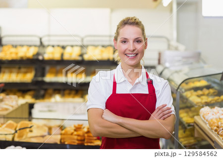 Woman in twenties wearing red apron standing behind counter in bakery with bread racks, glass case Woman in twenties wearing red apron standing behind counter in bakery with bread racks, glass case 129584562