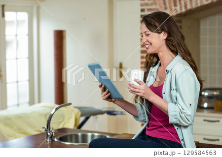 Woman sitting on kitchen island countertop holding light blue tablet and white mug, copy space Woman sitting on kitchen island countertop holding light blue tablet and white mug, copy space 129584563