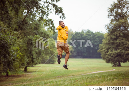 Man jumping wearing bright yellow hoodie and brown shorts twisting in grassy park along dirt path 129584566