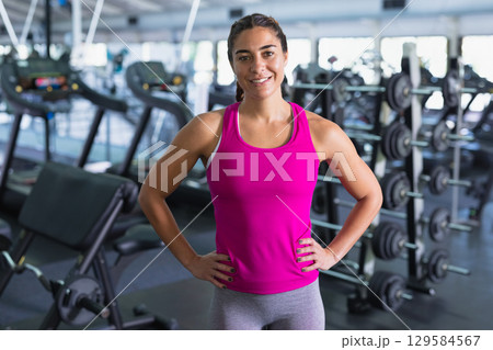 Woman standing confidently in modern gym wearing pink tank top by dumbbell rack and treadmills 129584567