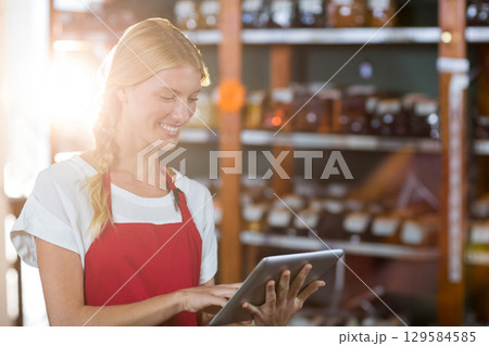 Female employee wearing red apron updating stock on tablet in aisle with shelves, jars, copy space Female employee wearing red apron updating stock on tablet in aisle with shelves, jars, copy space 129584585