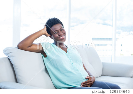 African American woman sitting on grey sofa cradling belly in modern living room with large windows 129584646