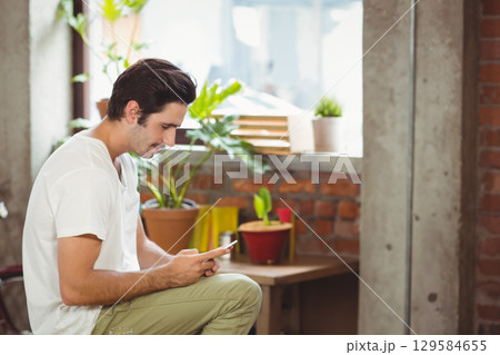 Man in mid-20s leaning forward examining smartphone on bench in loft with potted plants, copy space 129584655