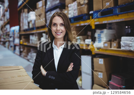Female warehouse manager in suit standing with arms crossed at aisle with metal shelving and boxes 129584656