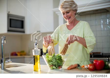 Senior woman tossing green salad in home kitchen using glass bowl and wooden servers, copy space 129584684