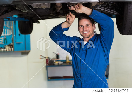 Male mechanic in coveralls smiling, holding wrench under lifted car in repair shop 129584701