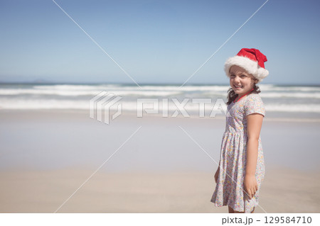 Female child standing at sandy beach, wearing red Santa hat and floral dress, copy space 129584710