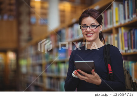 Mid adult woman standing among wooden bookshelves holding tablet and wearing backpack, copy space 129584718