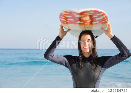 Woman standing waist-deep at shoreline in dark wetsuit holding red-and-white surfboard overhead Woman standing waist-deep at shoreline in dark wetsuit holding red-and-white surfboard overhead 129584725