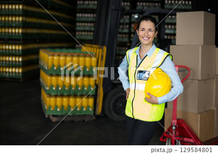 Female warehouse worker wearing safety vest holding hard hat amid beverage crates, copy space Female warehouse worker wearing safety vest holding hard hat amid beverage crates, copy space 129584801