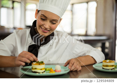 Female chef wearing white uniform at pastry station plating layered desserts on pale blue plates 129584809