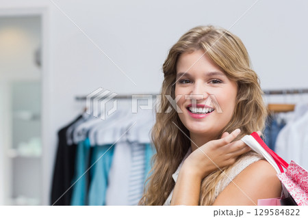 Female shopper in twenties smiling and holding pink shopping bags at shop with clothing rack 129584822