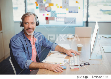 Mid-50s man wearing headphones sitting at desk in office facing monitor typing on wireless keyboard Mid-50s man wearing headphones sitting at desk in office facing monitor typing on wireless keyboard 129584826