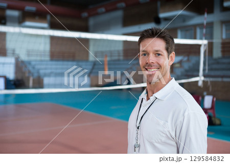Male coach standing near volleyball net on gymnasium court holding whistle on lanyard, copy space 129584832