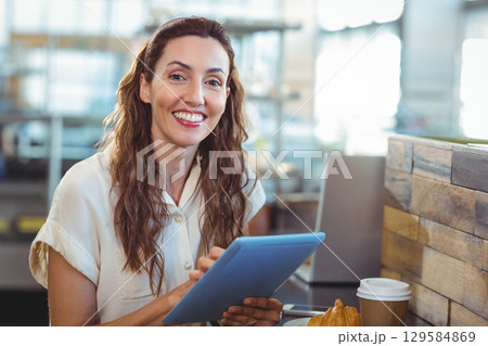 Woman smiling and holding tablet at cafe counter with coffee cup, croissant, copy space 129584869