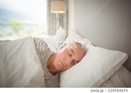 Senior man sleeping under white duvet in bedroom beside sliding glass door near balcony railing 129584874