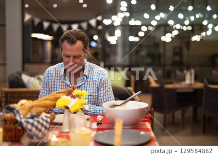 Senior man sitting at cafe dining table praying before meal with wicker bread basket 129584882